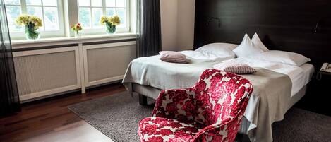 A hotel room with a bed, a chair, a window with flowers, and a radiator.