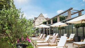 Eine Dachterrasse mit Liegestühlen und Sonnenschirmen, einer Wasserspende und einem Gebäude im Hintergrund mit traditioneller Architektur.