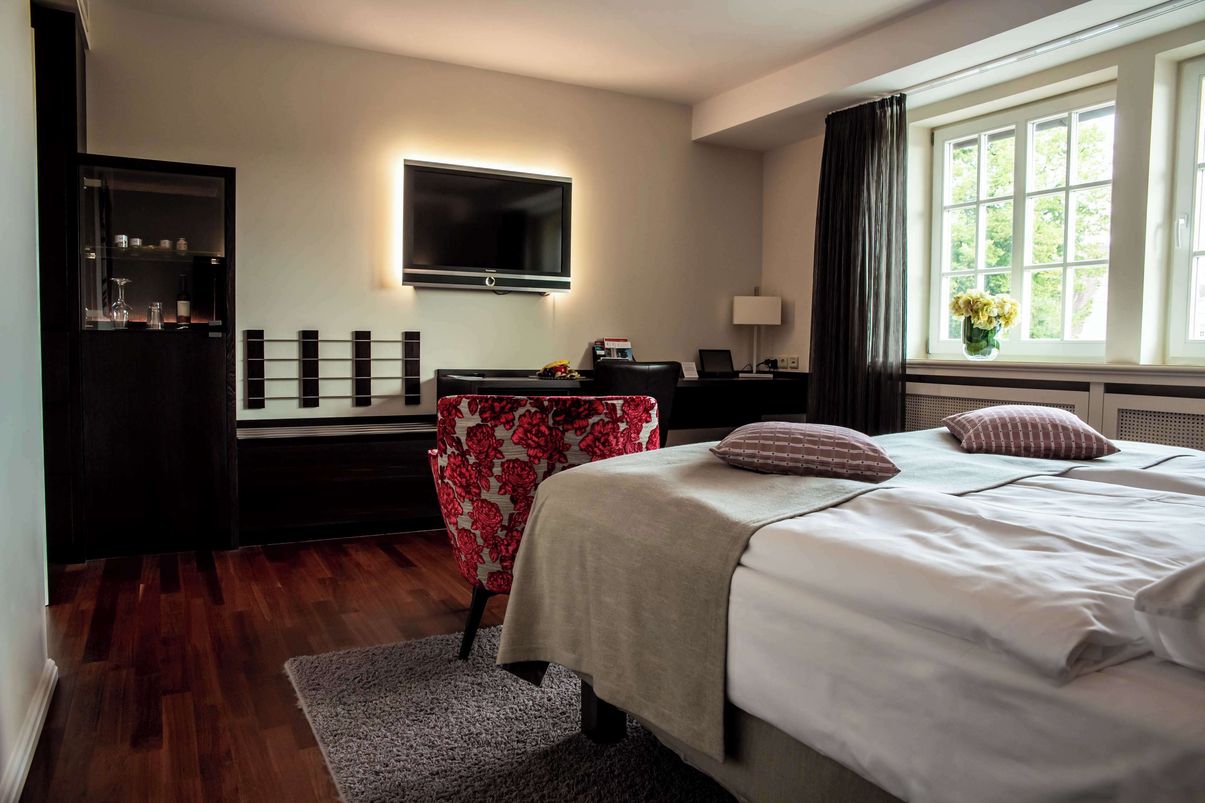 A hotel room with a bed, a desk with a computer, a TV mounted on the wall, and a window with curtains.