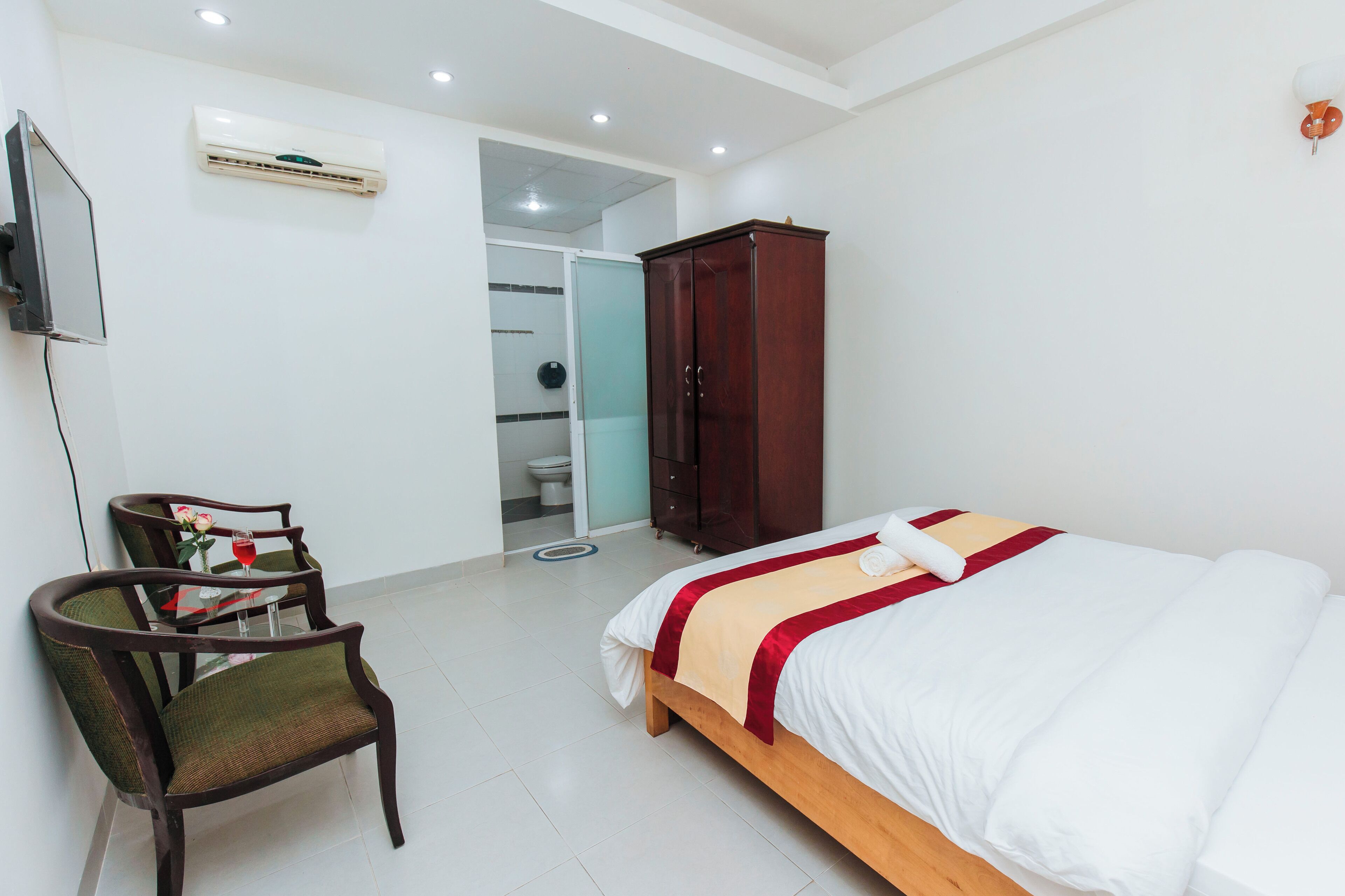 A hotel room with a bed, a wooden wardrobe, a chair, a small table with a glass top, and an air conditioning unit on the wall.