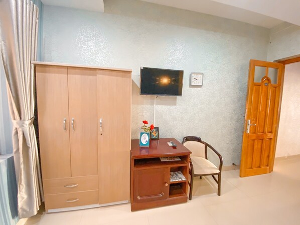 A room with a wooden wardrobe, a desk with a chair, a television mounted on the wall, and a wooden door.