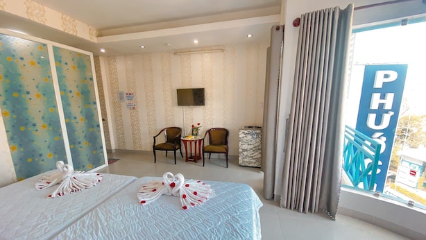 A hotel room with a large bed, two chairs, a small table, a TV, and a view of the outside. - Phuoc Hai Hotel By The Moment (Ho Chi Minh City)