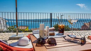 Een houten tafel met een hoed, zonnebril, een martini glas en een drankje op een balkon met uitzicht op zee.
