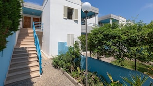 A modern building with a staircase leading to a balcony, surrounded by greenery and a blue wall.