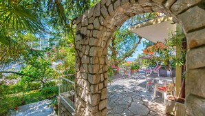 A stone archway leading to a patio area with outdoor seating and a view of a residential building. - Holiday apartment with balcony and sea view (Selce)