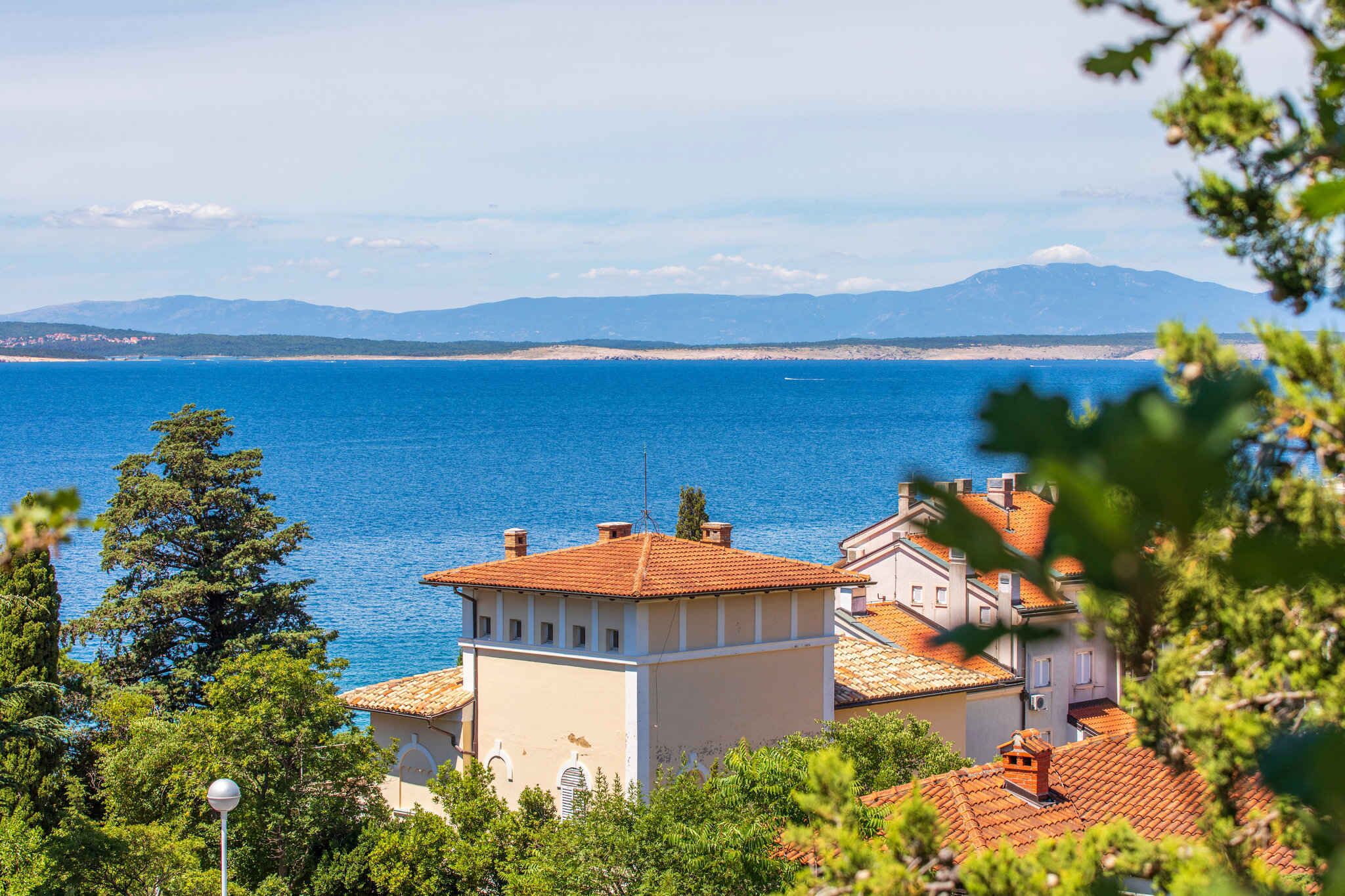A coastal building with a red-tiled roof, surrounded by greenery, overlooking a calm sea and distant mountains.
