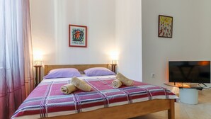 Cama bem arrumada com manta listrada, duas almofadas e toalhas enroladas. Há duas luminárias de cabeceira, uma TV em um suporte e duas obras de arte na parede.