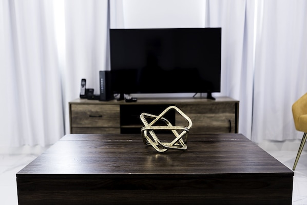 Une table en bois foncé, ornée d’une sculpture métallique aux motifs géométriques au centre, un téléviseur posé sur un meuble en bois à l’arrière-plan, et une chaise jaune sur la droite.