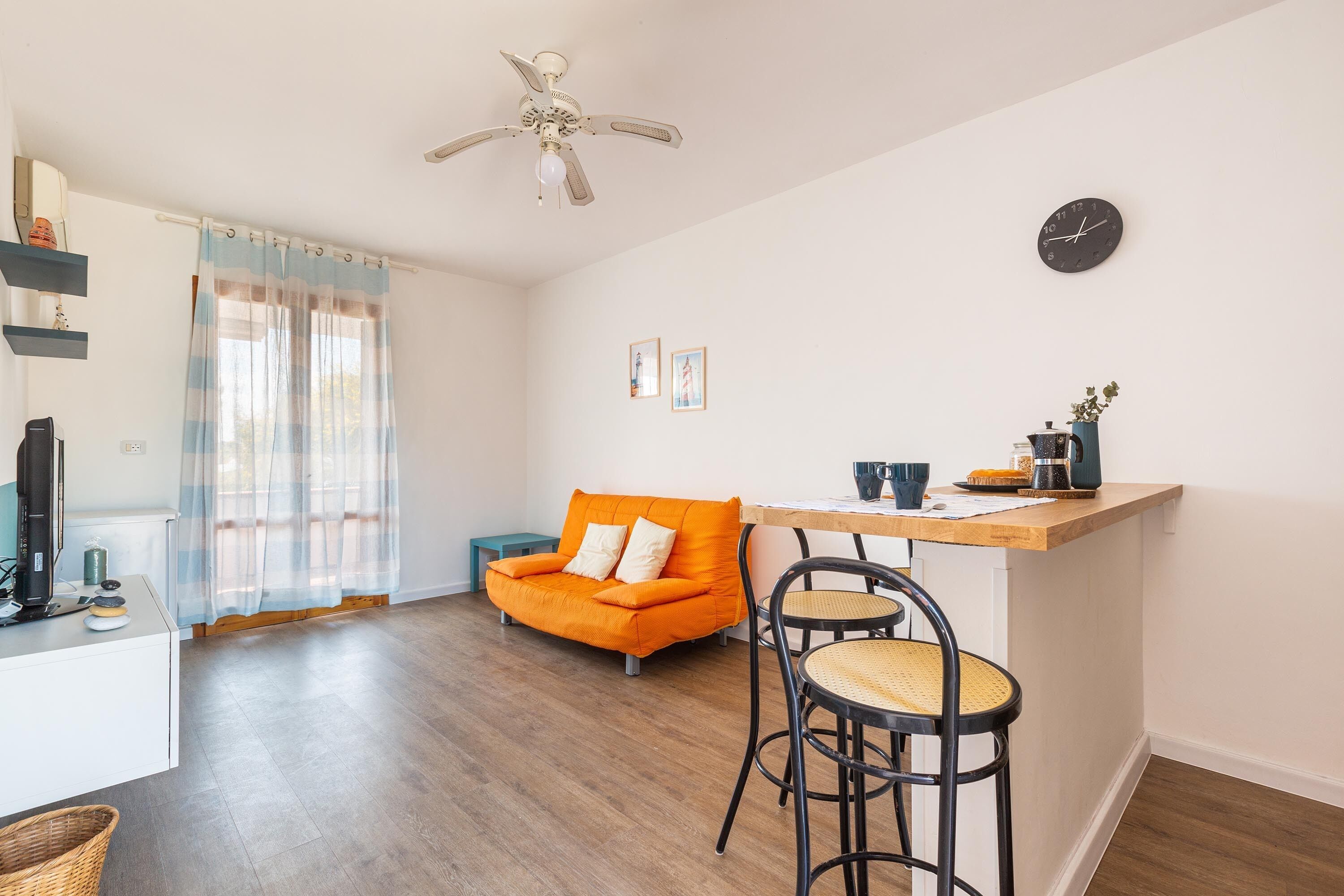 A modern living space with a sofa, a dining area with a bar, a ceiling fan, and a wall clock.