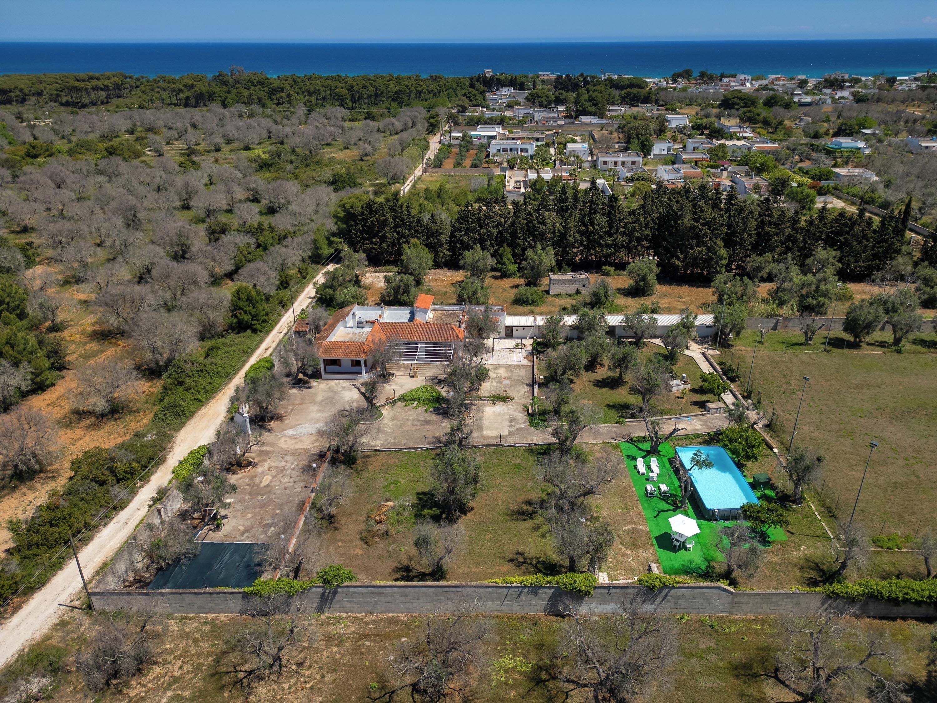 An aerial view of a residential area with a large house featuring a swimming pool, surrounded by trees and a clear view of the sea.