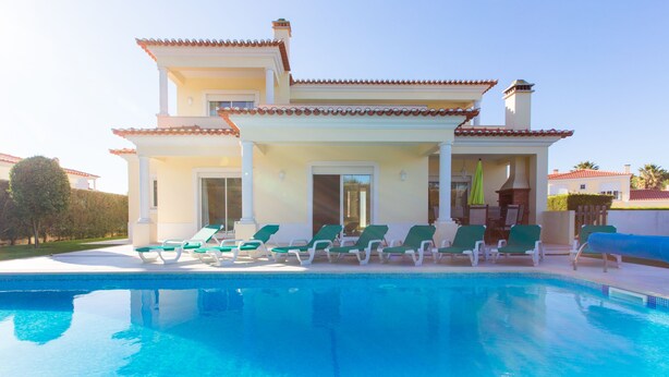 Une maison à deux étages avec une piscine, des fauteuils de jardin verts et un patio couvert.