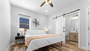 A bedroom with a bed, bedside tables, a ceiling fan, a window, and a bathroom visible through an open door. - Lake Views Hot Tub Sauna Arcade Decks Family Stay (Branson)
