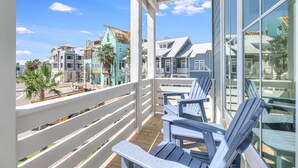Ein Balkon mit Holzdeck und weißen Geländern, der auf eine Küstenregion mit bunten Gebäuden und Palmen blickt.