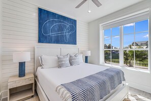Une chambre avec un grand lit, deux tables de chevet avec des lampes, un tableau abstrait bleu et une grande fenêtre donnant sur des maisons et de la verdure.