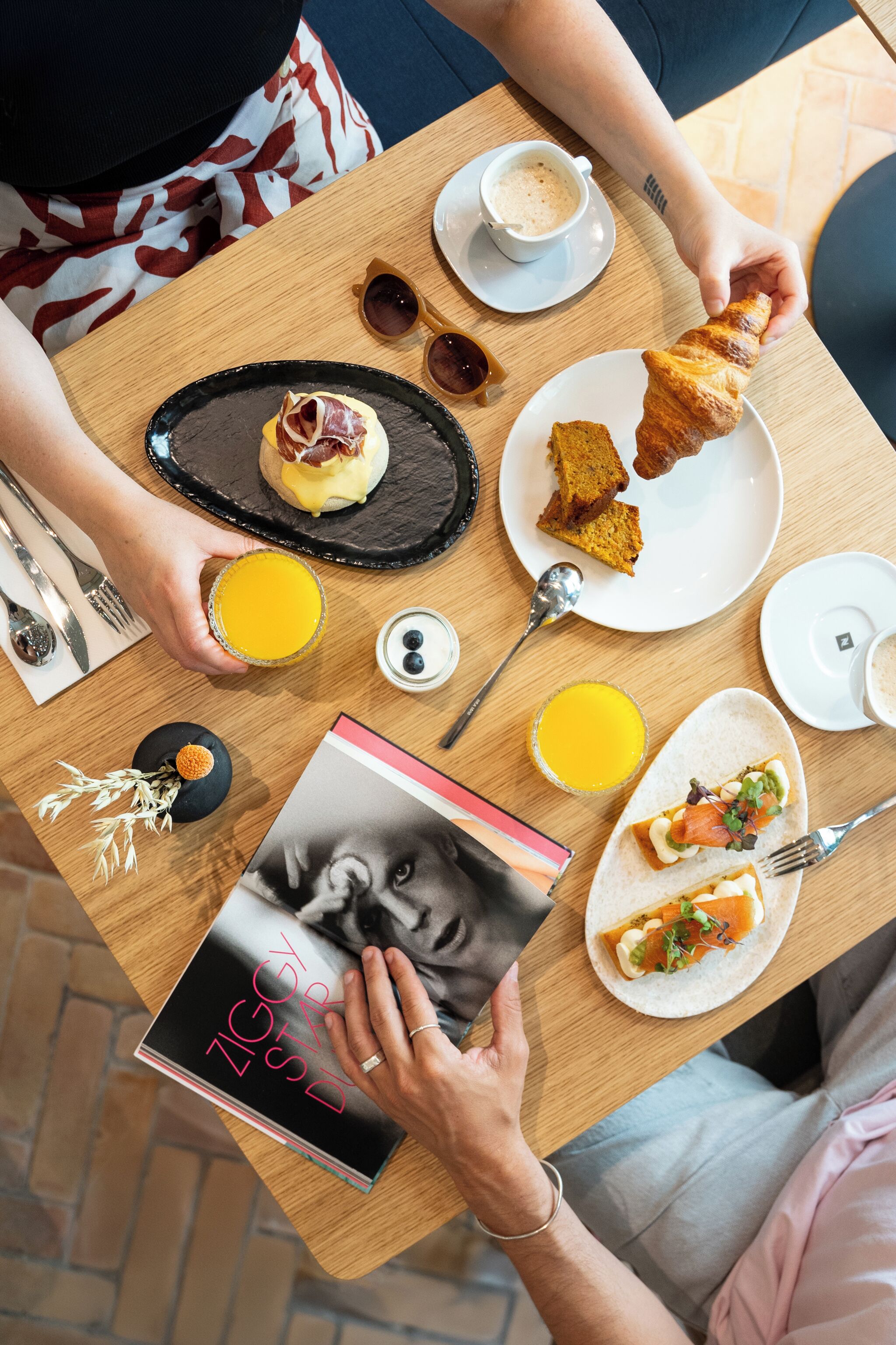 Une table de petit-déjeuner avec des croissants, un sandwich aux croissants, une assiette de nourriture, un verre de jus d’orange, une tasse de café et un magazine.