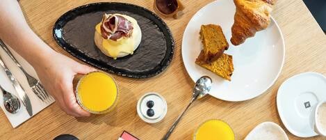 Una mesa de desayuno con croissants, un sándwich de croissant, un plato de comida, un vaso de jugo de naranja, una taza de café y una revista.
