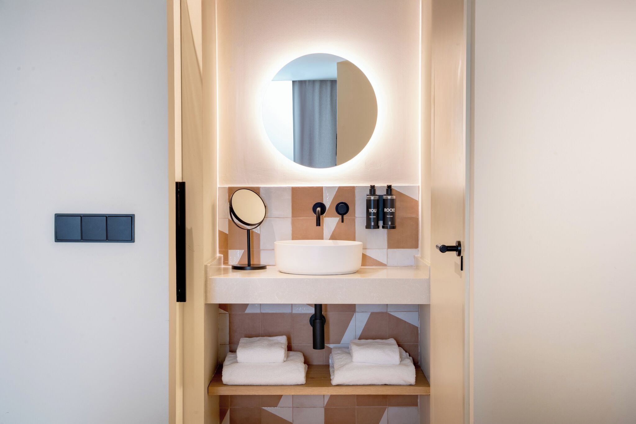 A modern bathroom with a round mirror, a white sink, and two shelves with folded towels.