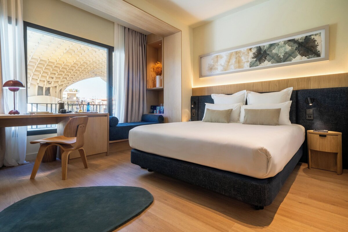 A modern hotel room with a large bed, a desk, a chair, a small side table, and a window with a view of a building.