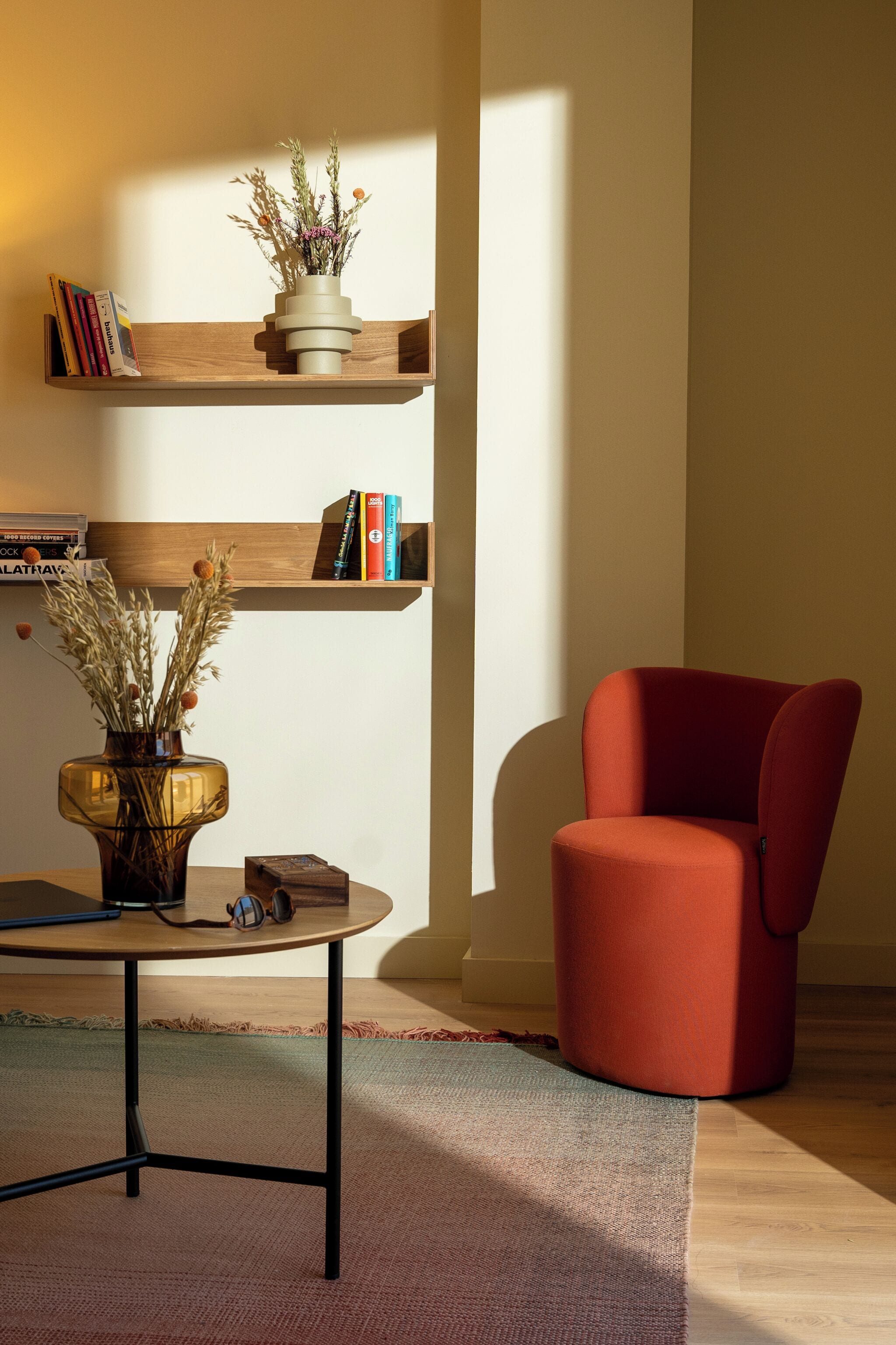 Een moderne woonkamer met een rode fauteuil, een houten plank met boeken, een vaas met gedroogde planten, en een kleine tafel met een laptop en hoofdtelefoon.