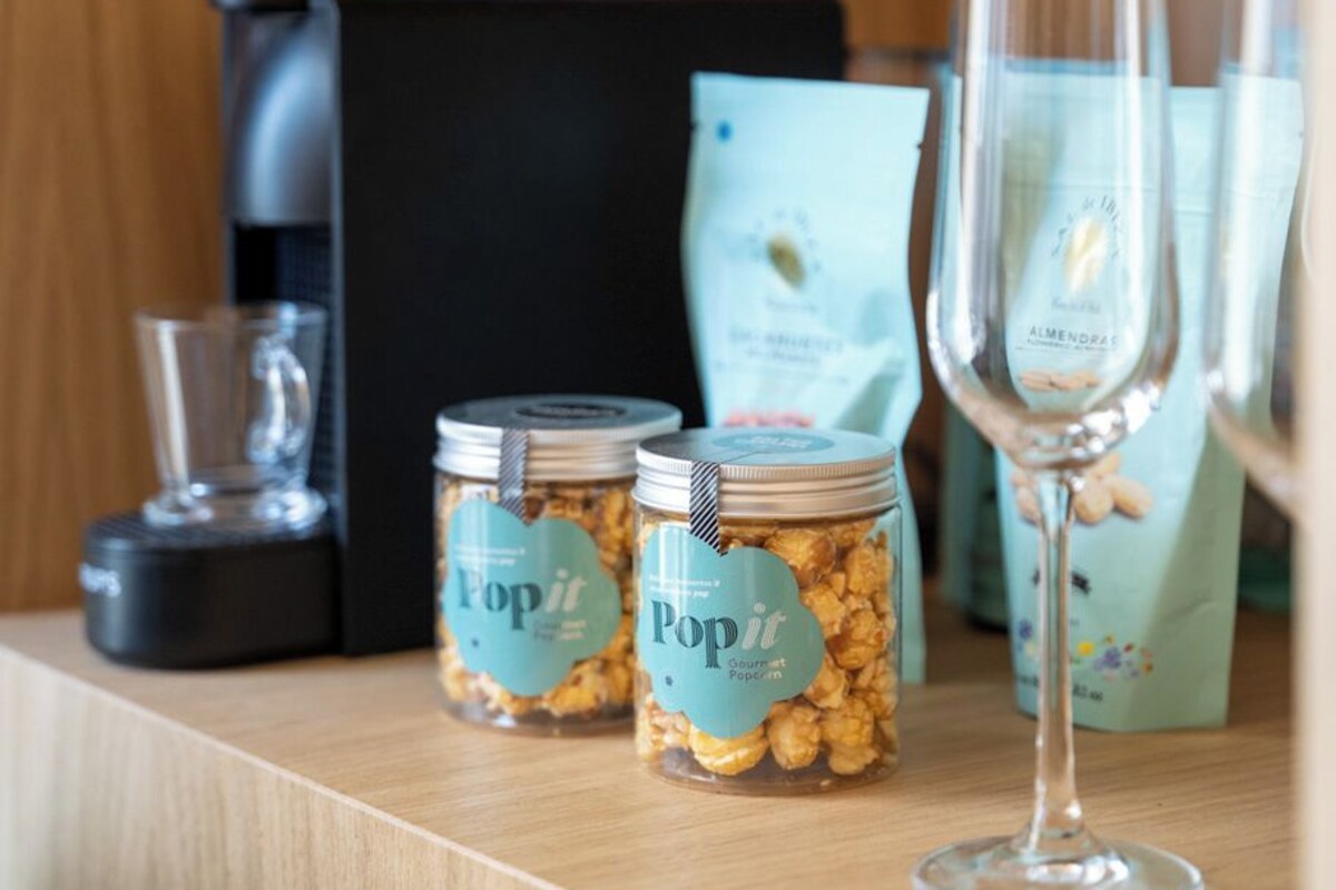 Dos frascos de Popit Gourmet Popcorn, una copa de vino de vidrio y una máquina de café negra sobre una superficie de madera.