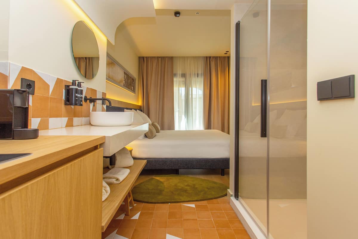 A modern hotel room with a wooden vanity, a large bed, a shower area, and a view of the outdoors.