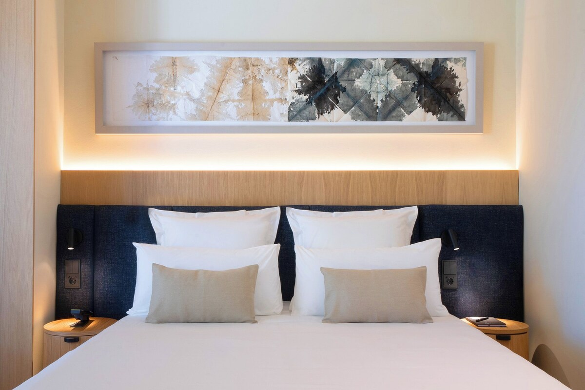 A neatly made double bed with white and beige pillows, a dark headboard, and a framed abstract painting above.