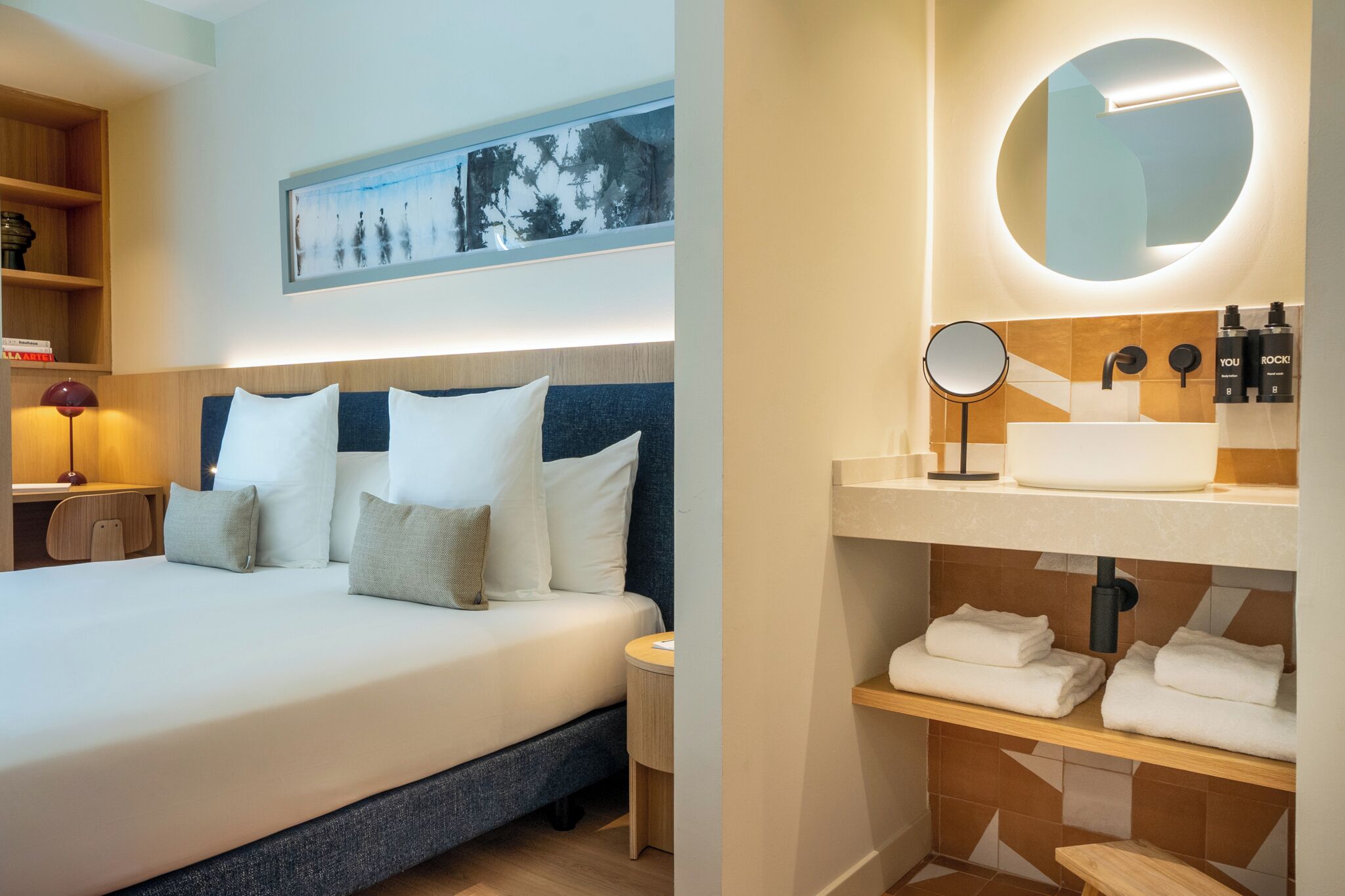 A hotel room with a bed, a bedside table with a lamp, a wall-mounted picture, and a bathroom with a sink, mirror, and towels.