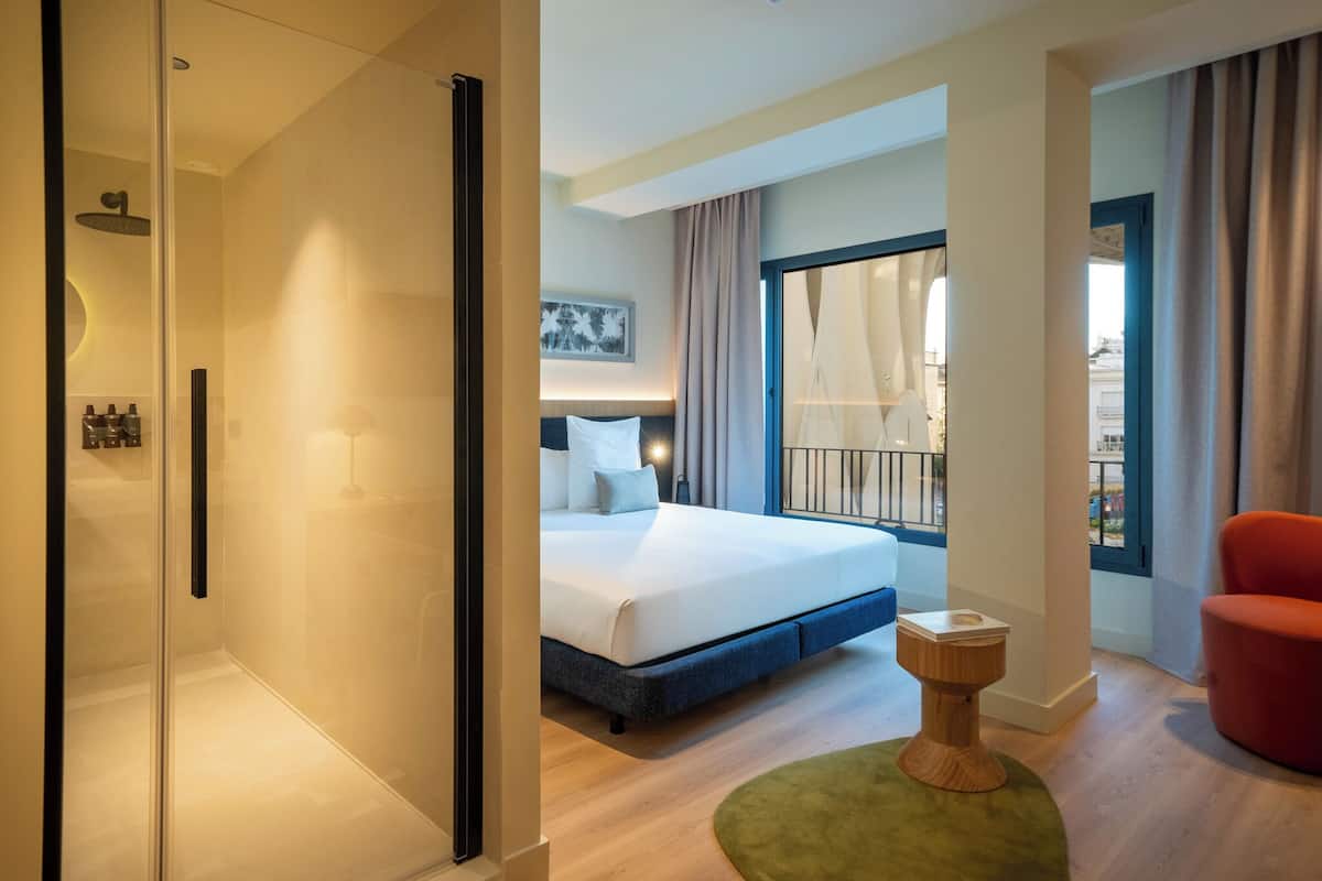 A modern hotel room with a glass-enclosed shower, a bed with a headboard, a small wooden side table, and a red chair.