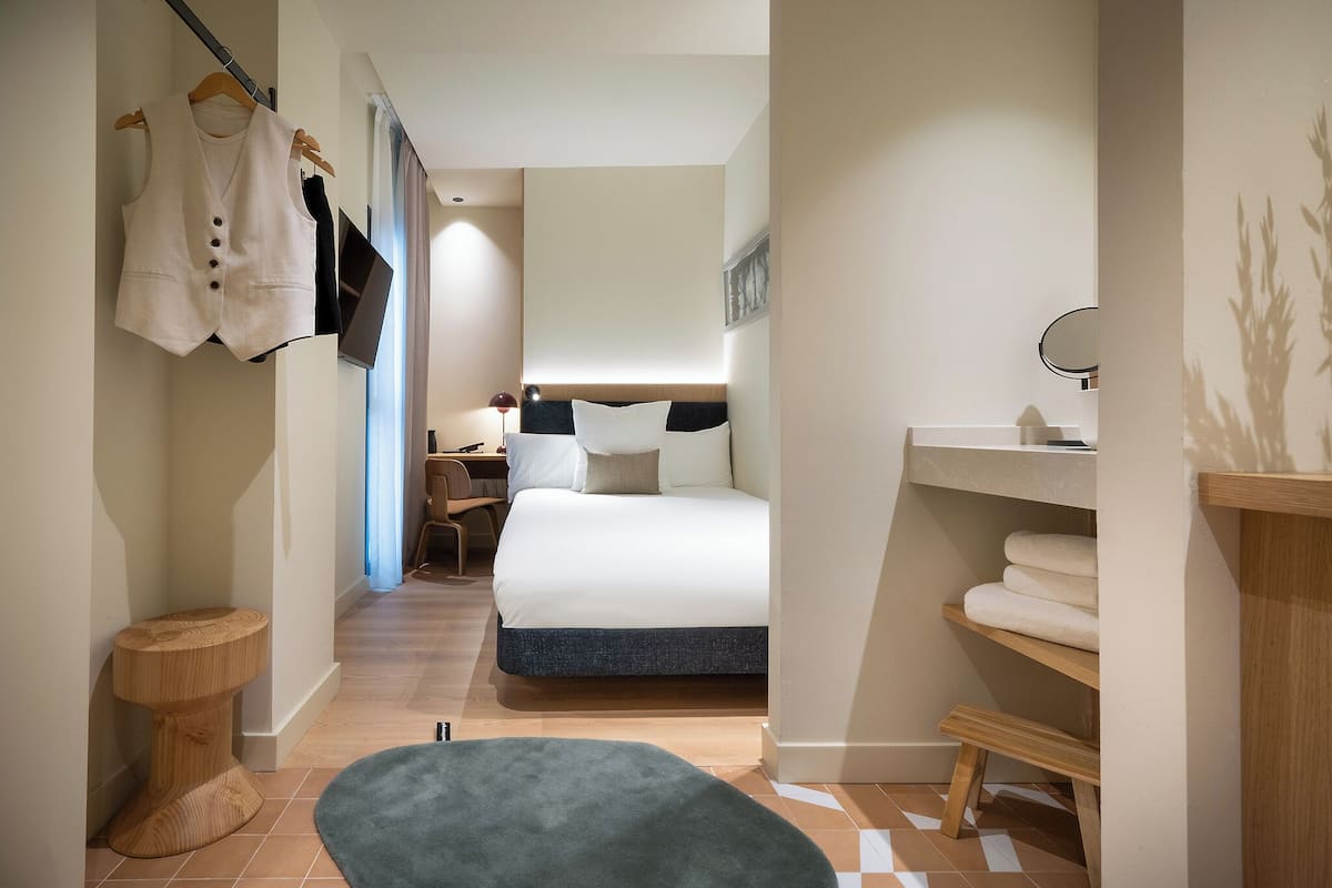 A modern hotel room with a bed, a desk, a chair, a small round table, a hanging coat rack, and a shelf with towels.