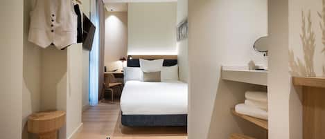 A modern hotel room with a bed, a desk, a chair, a small round table, a hanging coat rack, and a shelf with towels.