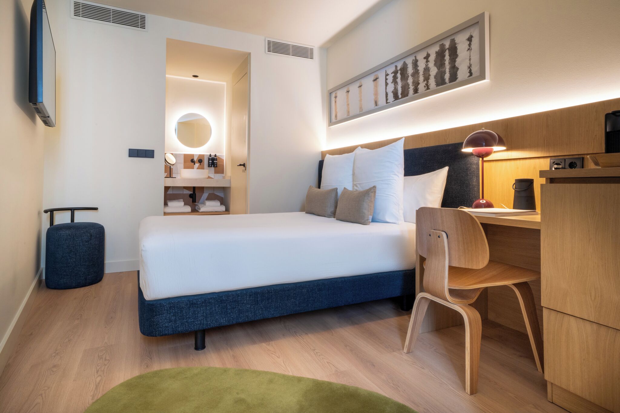 A modern hotel room with a bed, desk, chair, and a small kitchenette.