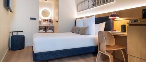 A modern hotel room with a bed, desk, chair, and a small kitchenette.