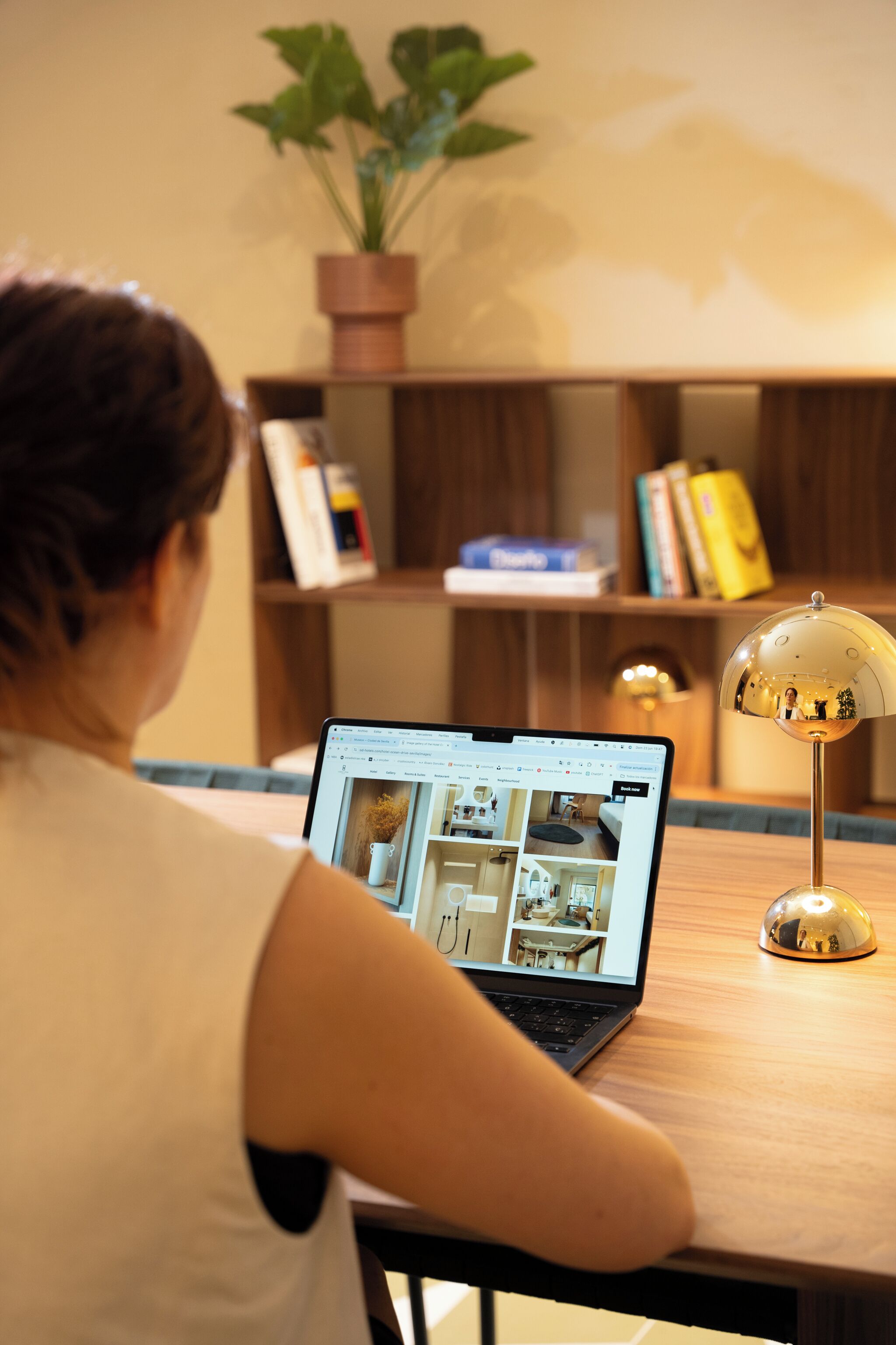 A person sitting at a wooden table with a laptop displaying a webpage. There is a potted plant and a lamp on the table.