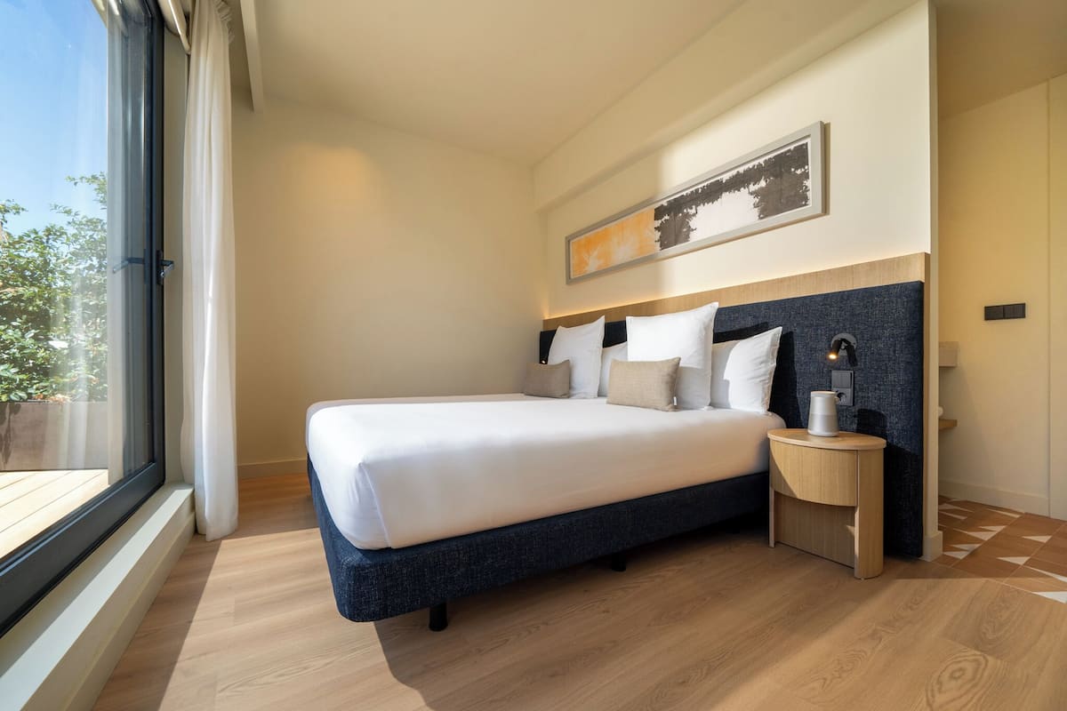 A modern hotel room with a large bed, a bedside table with a lamp, and a window with curtains.