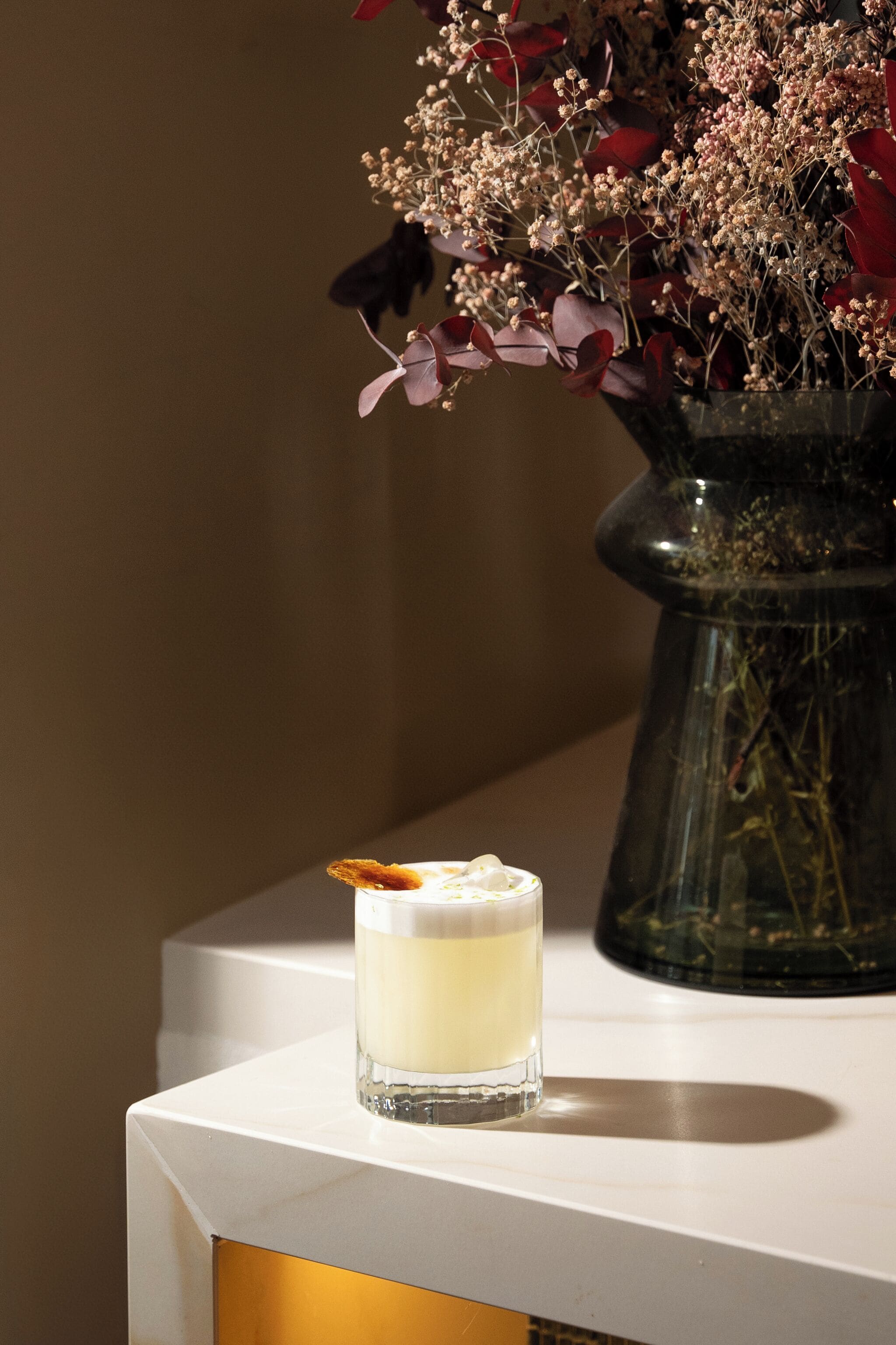 A glass of a creamy beverage with a garnish on a white surface, next to a vase with dried flowers.