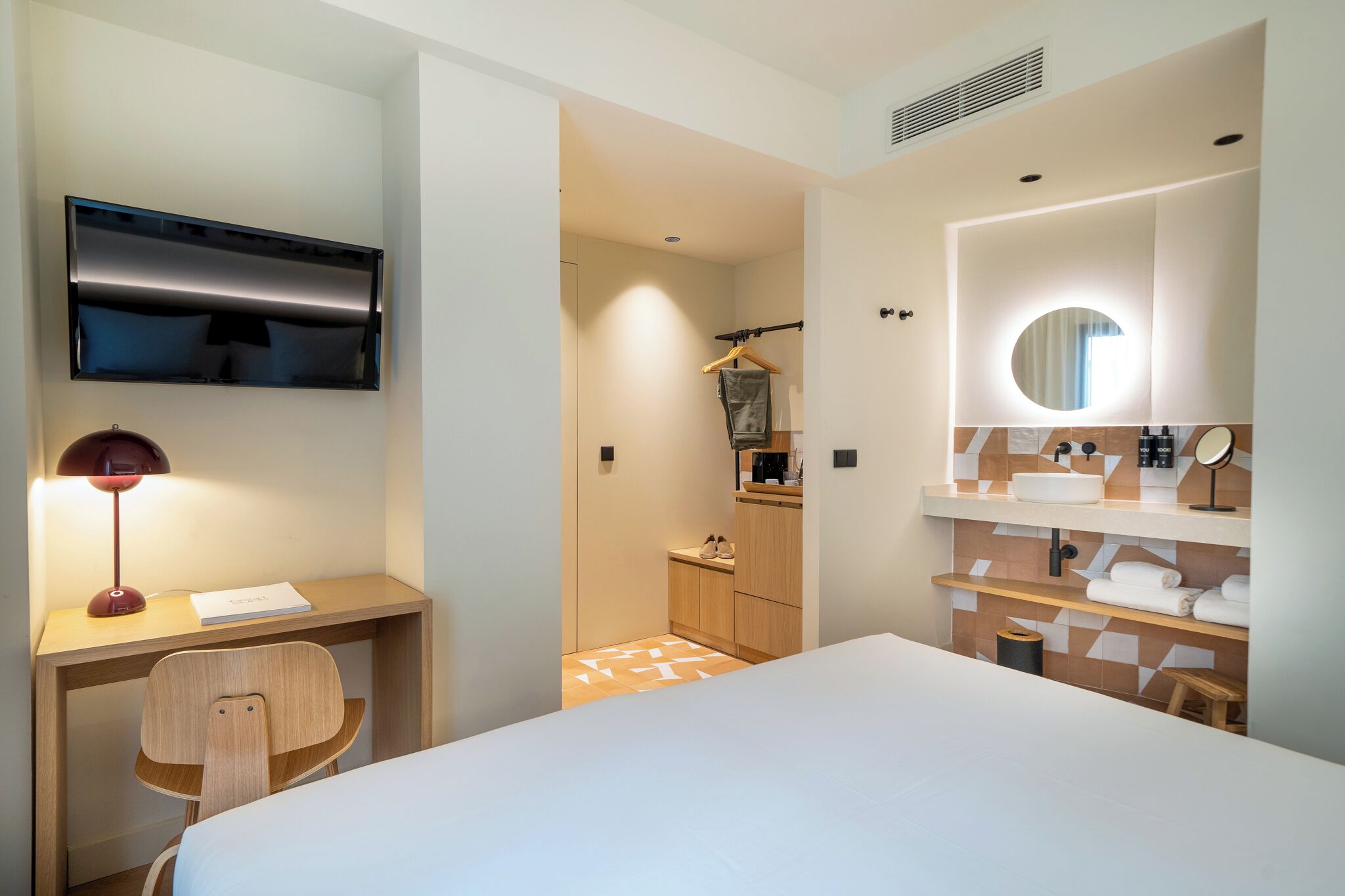 A hotel room with a bed, desk, chair, TV, and a bathroom with a mirror, sink, and towel rack.