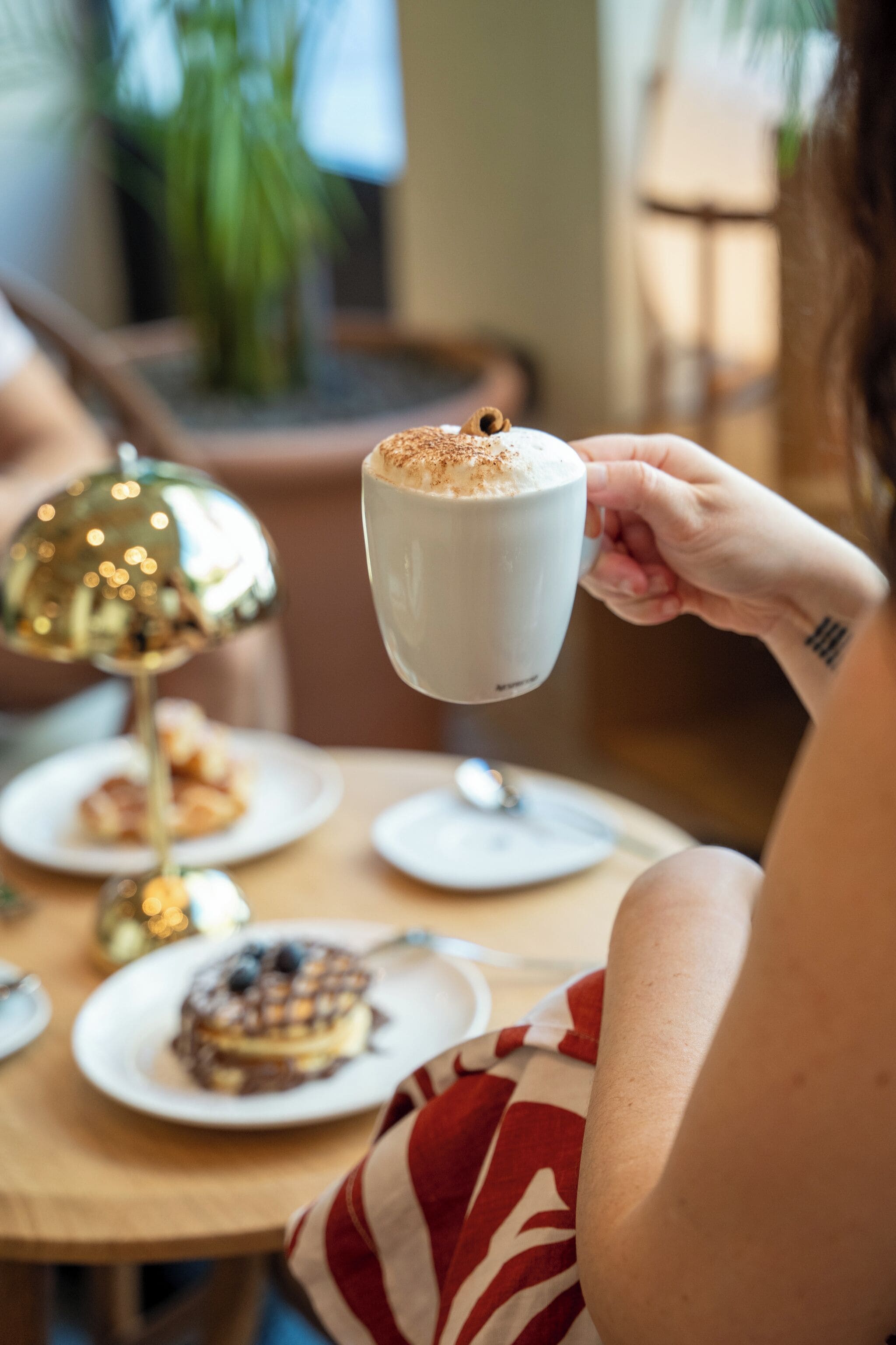 A person holding a cup of coffee with a frothy top, a plate with a pastry, and a decorative lamp on a table.