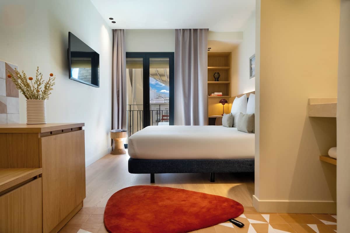 A modern hotel room with a bed, a wooden desk, a red rug, and a view of the city through the balcony doors.