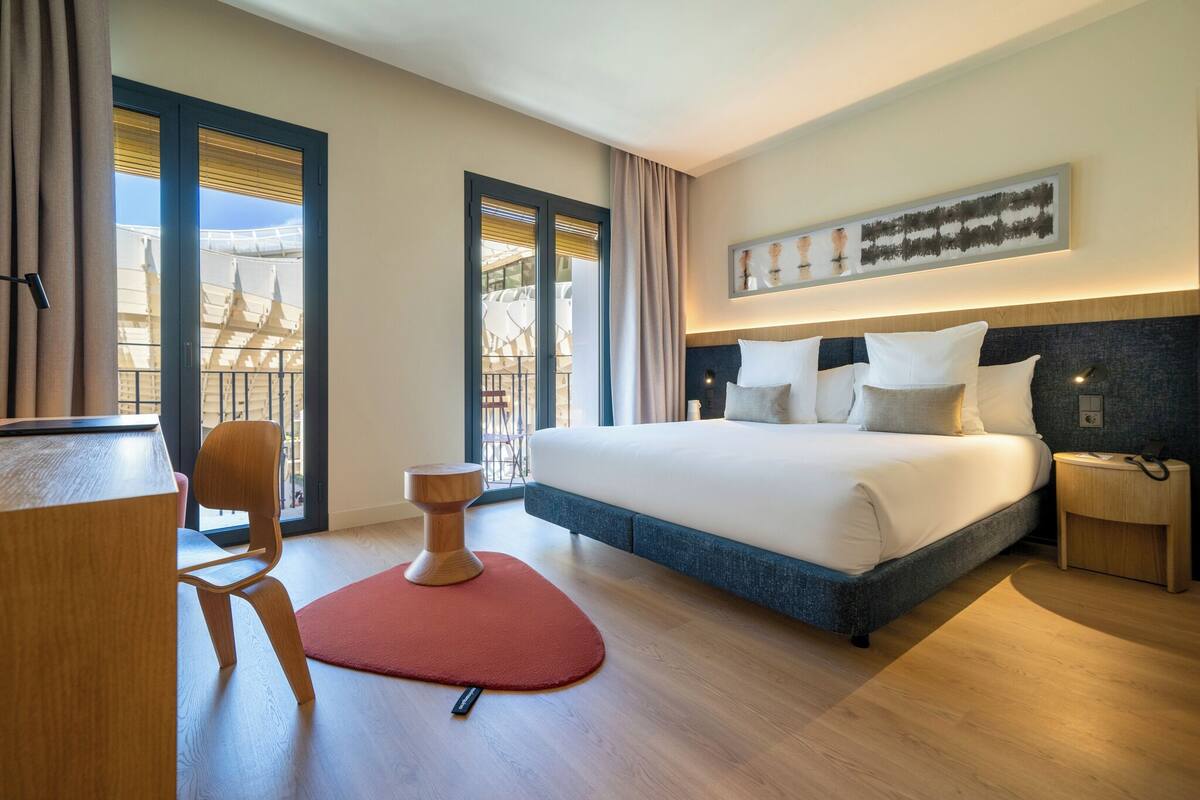 A modern hotel room with a large bed, a desk, and a chair. There is a balcony with a view of buildings.