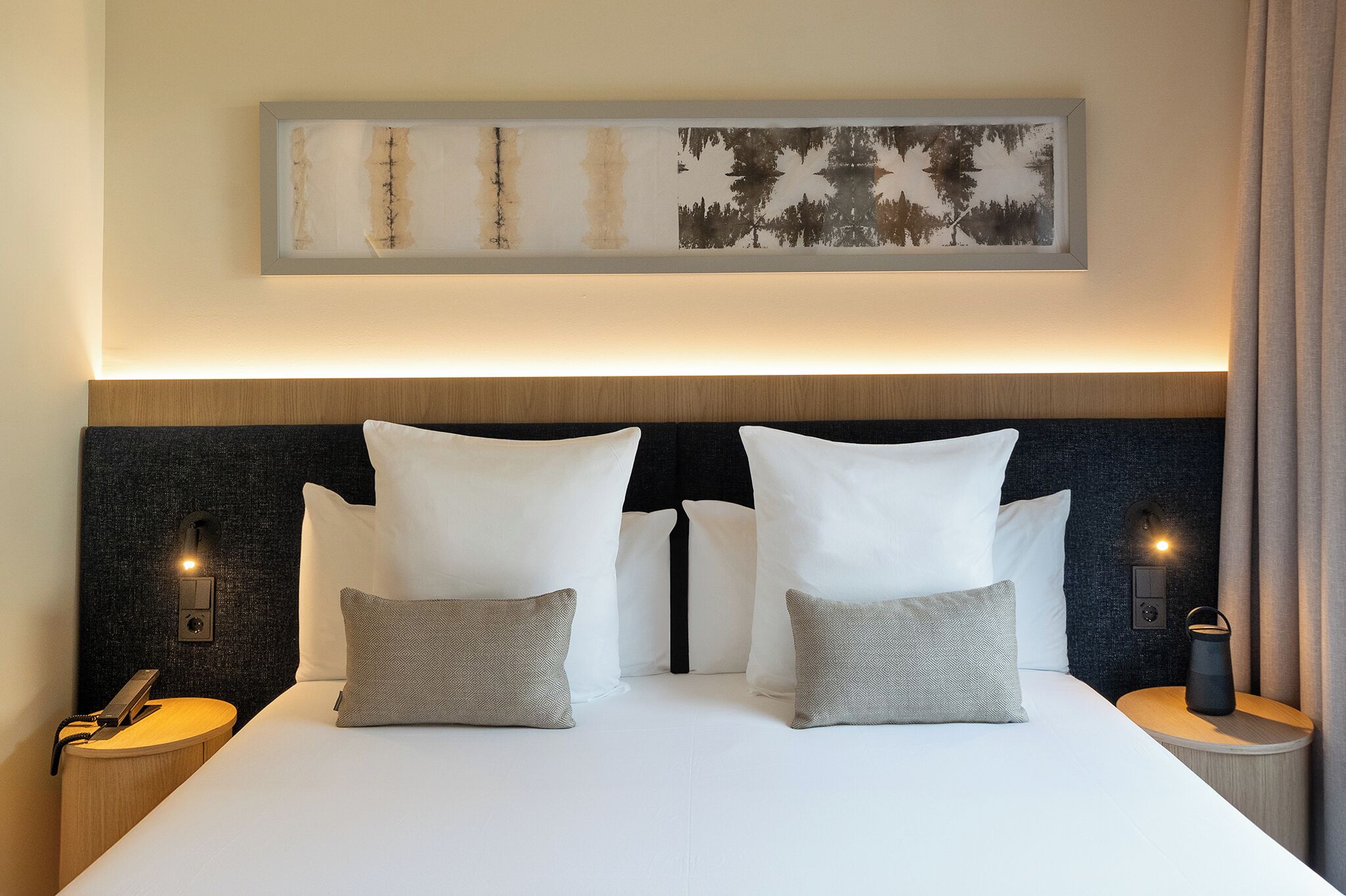Una cama bien hecha con almohadas blancas y grises, un cabecero oscuro con textura y dos lámparas de noche.