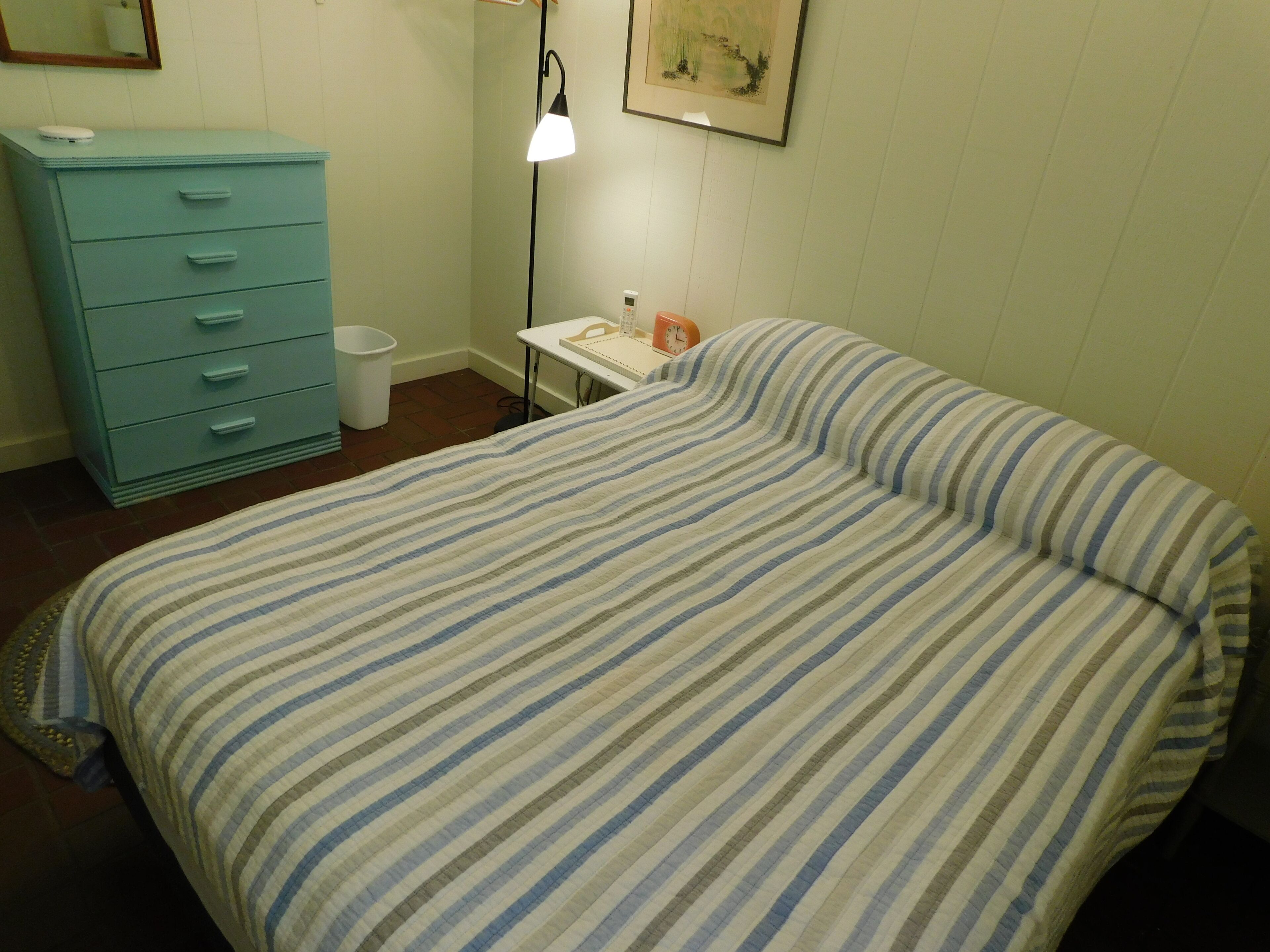 A bedroom with a striped bedspread, a blue dresser, a white bedside table with a lamp, and a trash bin.