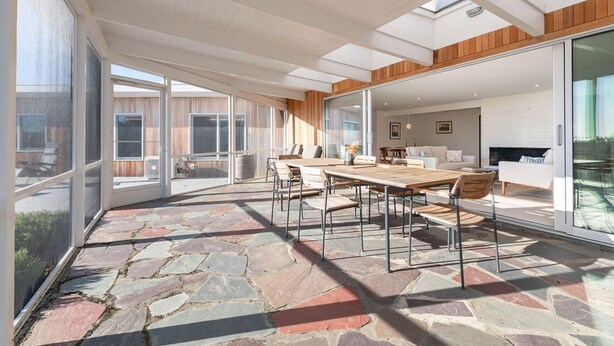A spacious patio area with a stone floor, a wooden table, and chairs, overlooking a modern interior with a fireplace. - Newly Reno'd GORGEOUS, Water View, Beach Access (CS0034) (Wellfleet)
