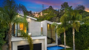 A modern two-story house with a swimming pool, surrounded by lush greenery and palm trees.