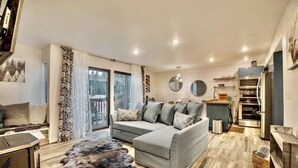 A modern living room with a grey sectional sofa, a fur rug, and a kitchen area with a stove and cabinets. - Updated tree top cabin retreat! (Mammoth Lakes)