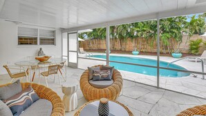 Une terrasse extérieure moderne, dotée d’une piscine protégée par des parois en verre, de meubles en osier et offrant une vue sur un jardin aménagé.