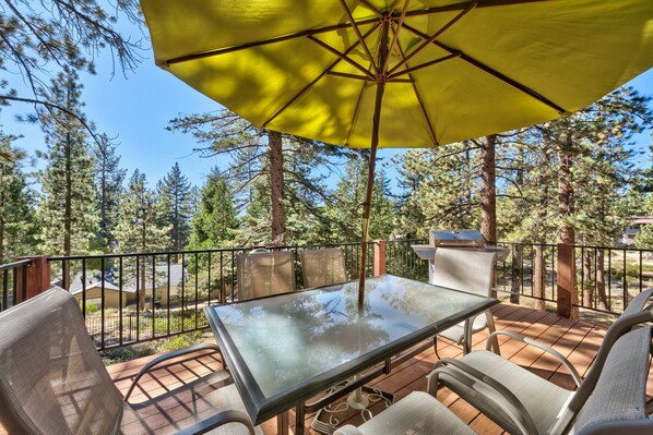 An outdoor patio with a glass table and chairs, a yellow umbrella, and a grill.