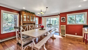 Comedor con mesa de madera, sillas y bancos blancos. Se observa un aparador de madera con puertas de cristal y una lámpara colgante sobre la mesa.