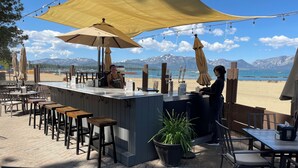 Un bar situé en bord de mer, avec une terrasse, un comptoir et une vue sur la plage et les montagnes.
