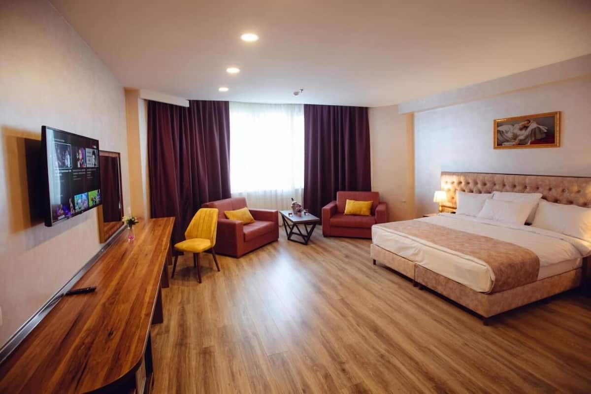 A hotel room with a bed, two armchairs, a small table, a TV mounted on the wall, and a window with curtains.