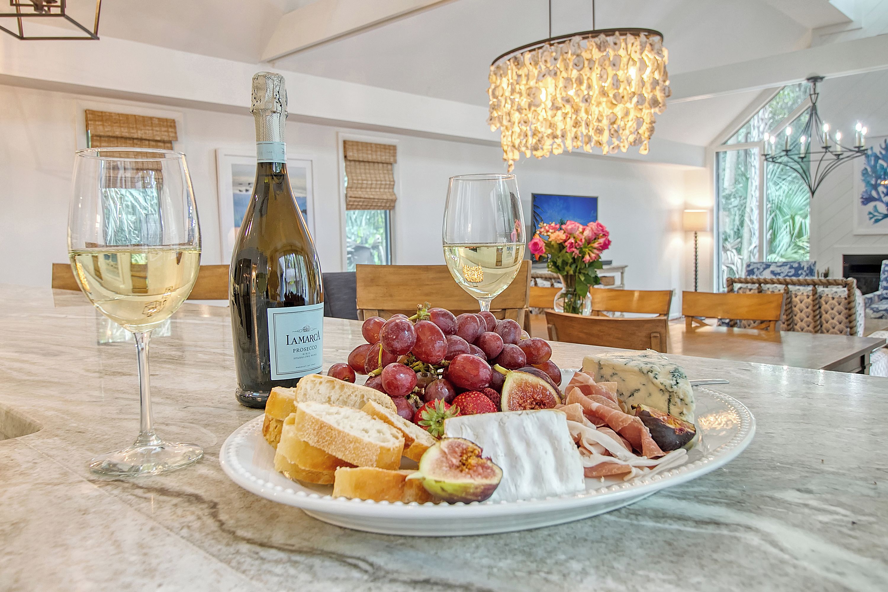 A dining table with a bottle of wine, two glasses of white wine, a plate of cheese, grapes, and bread.